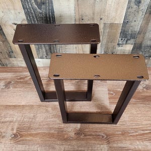 May include: Two metal table legs with a V-shaped design. The legs are a dark bronze color and have a rectangular top with pre-drilled holes for mounting. The legs are designed for furniture construction.