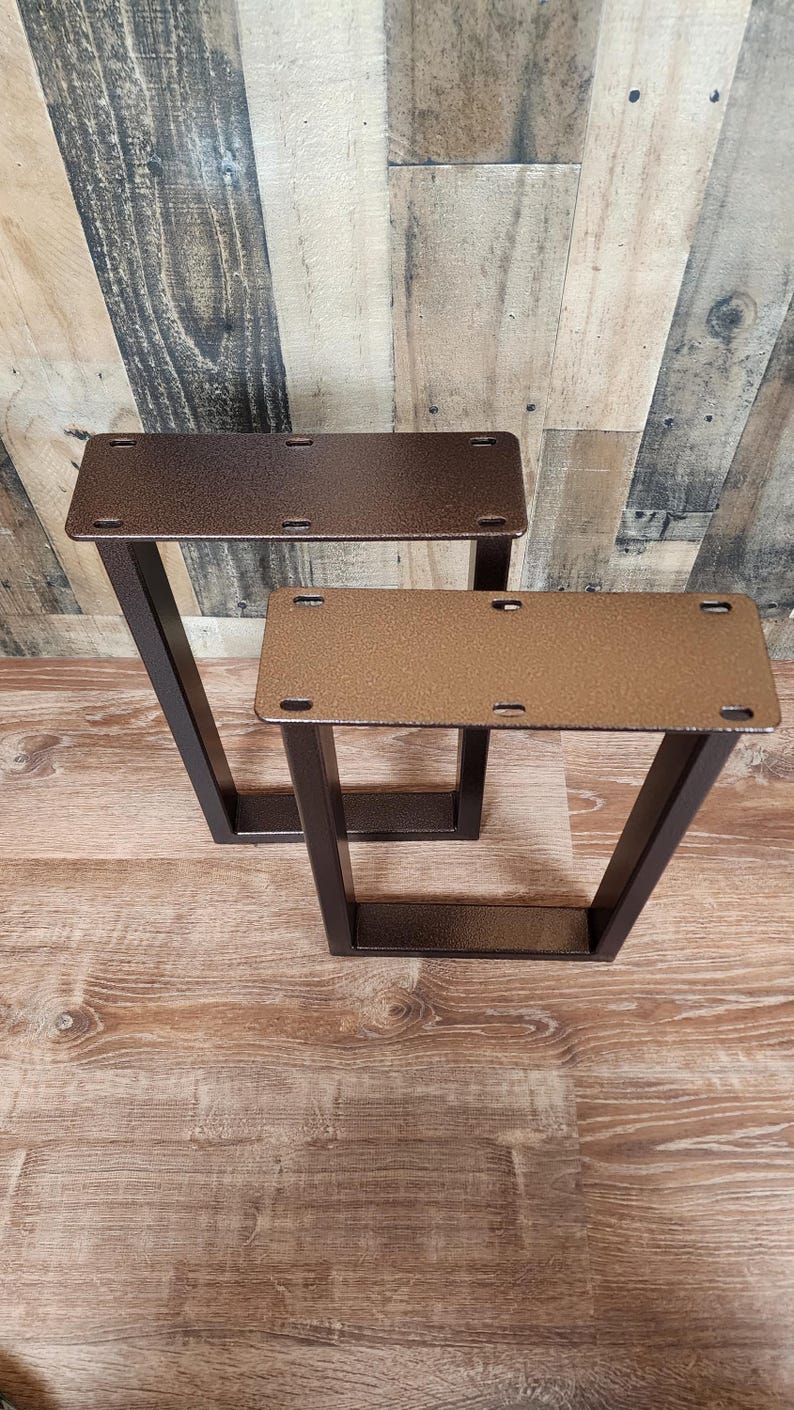 May include: Two metal table legs with a V-shaped design. The legs are a dark bronze color and have a rectangular top with pre-drilled holes for mounting. The legs are designed for furniture construction.