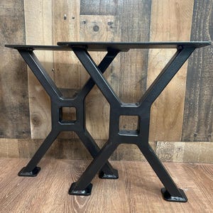 May include: Two black metal table legs with an X-shaped design. Each leg has a square base and a flat top with pre-drilled holes for mounting. The legs are made of sturdy metal and are suitable for various table projects.