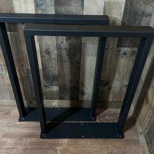 May include: Two black metal table legs with a modern, geometric design. Each leg has a rectangular top and base, connected by angled supports. The legs are made of sturdy metal and are suitable for various table projects.