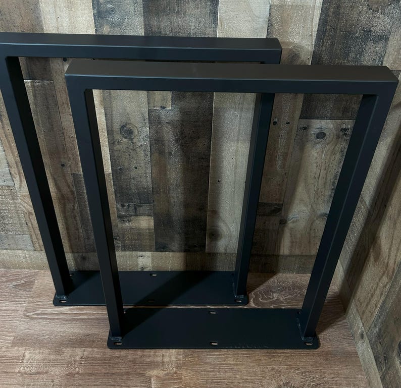 May include: Two black metal table legs with a modern, geometric design. Each leg has a rectangular top and base, connected by angled supports. The legs are made of sturdy metal and are suitable for various table projects.