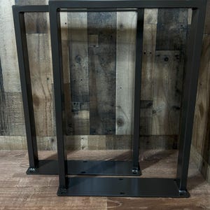 May include: Two dark gray metal table legs. Each leg has a rectangular frame design with a flat base and top. The legs are made of steel and are suitable for a variety of table tops. The legs are shown against a wood background.