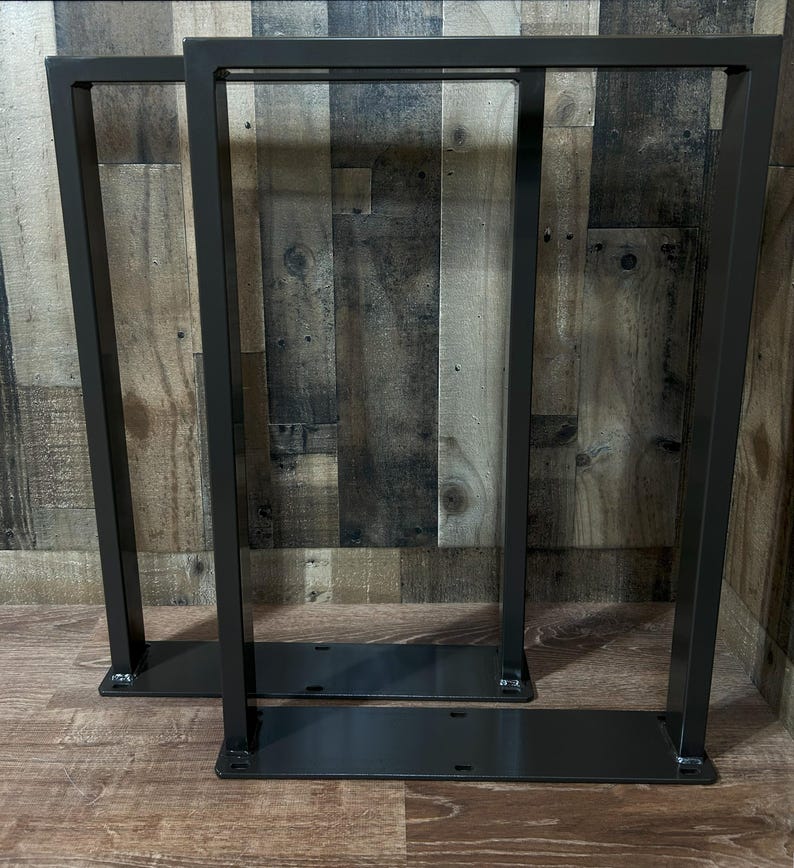 May include: Two dark gray metal table legs. Each leg has a rectangular frame design with a flat base and top. The legs are made of steel and are suitable for a variety of table tops. The legs are shown against a wood background.