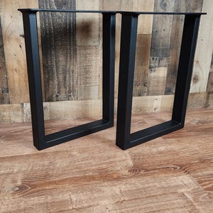 May include: Two black, square-tube steel table legs. The legs are U-shaped and have a matte finish. They are designed to support a tabletop. The legs are displayed on a wood-grain surface.