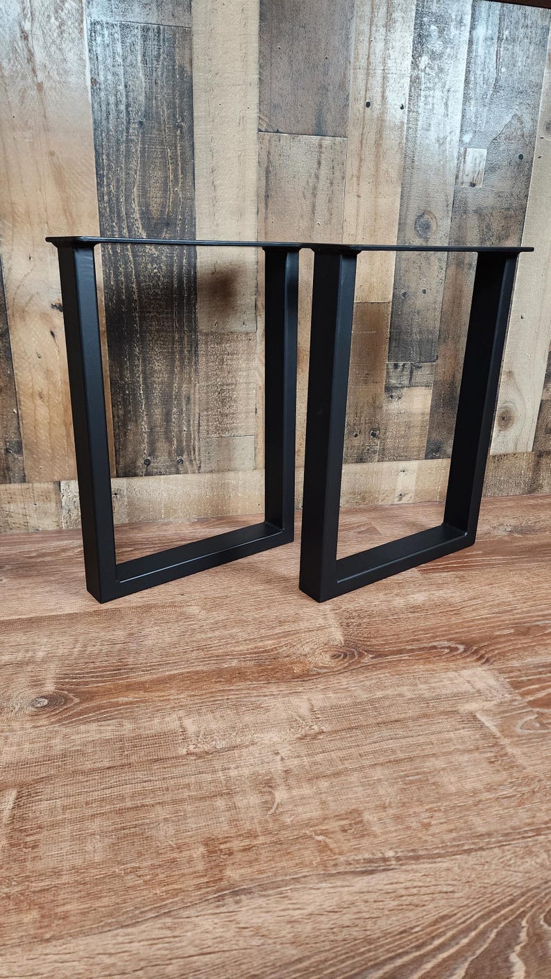 May include: Two black, square-tube steel table legs. The legs are U-shaped and have a matte finish. They are designed to support a tabletop. The legs are displayed on a wood-grain surface.