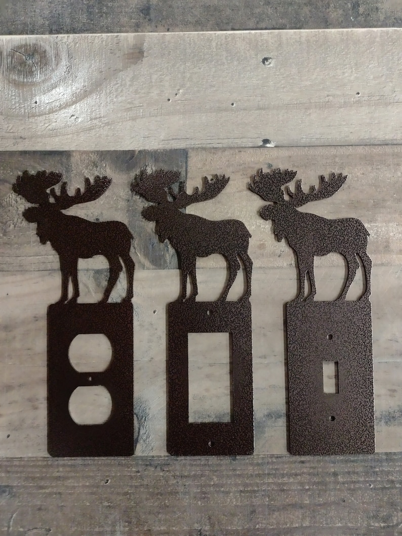 May include: Three metal moose silhouettes in a brown finish, each mounted on a different switch plate. The plates feature a double outlet, a single switch, and a light switch.