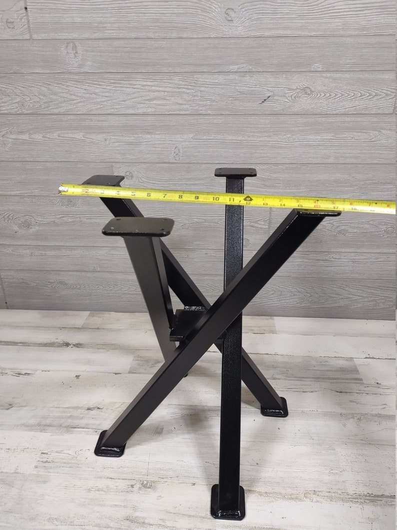 Op de afbeelding: Zwarte metalen tafelpoten met een X-vormig ontwerp. De poten zijn vierkant en meten ongeveer 30 cm breed. Een gele meetlint ligt over de bovenkant van de poten.