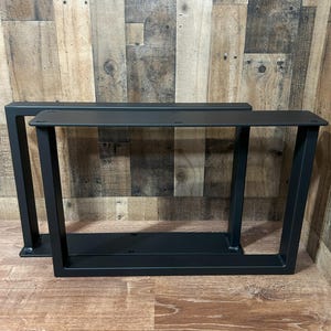 May include: Two black metal table legs with a modern, geometric design. Each leg features a rectangular top plate with pre-drilled holes for easy attachment. The legs have a sturdy, angled frame, perfect for a DIY project or furniture upgrade.