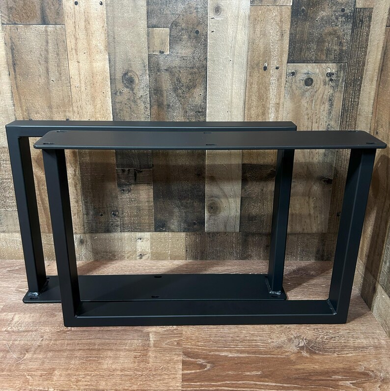May include: Two black metal table legs with a modern, geometric design. Each leg features a rectangular top plate with pre-drilled holes for easy attachment. The legs have a sturdy, angled frame, perfect for a DIY project or furniture upgrade.