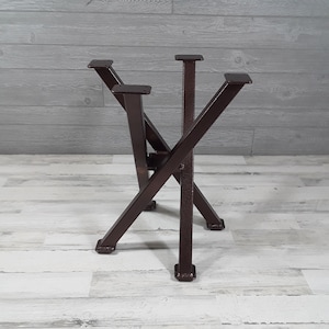 May include: A brown metal table base with an X-shaped design. The base has four legs and four mounting plates for attaching a tabletop. The metal has a textured finish, and the base is designed for stability and support.