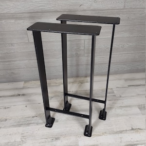 May include: Two black metal table legs with a rectangular top. The legs are made of square tubing and have a simple, industrial design.