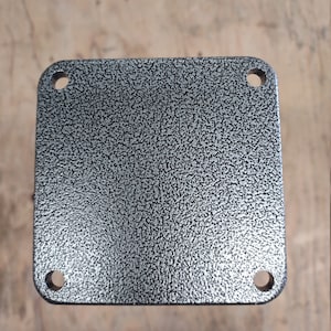 Puede incluir: Una placa cuadrada de metal con una superficie texturizada y cuatro agujeros alrededor de los bordes. La placa es de color plateado.