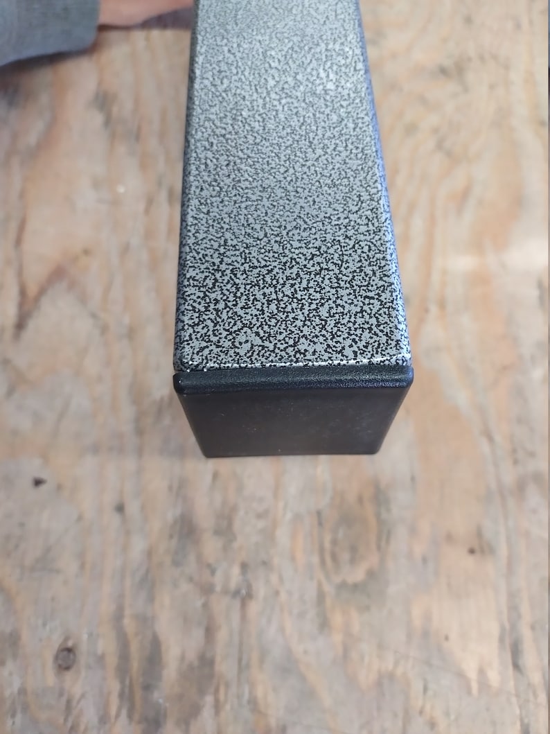 Puede incluir: Primer plano de una pata de mesa de metal gris con una tapa de pl&aacute;stico negro. El metal tiene un acabado texturizado y moteado.