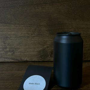 May include: A matte black can with a matching label that reads "Matte Black". The can is cylindrical and appears to be made of metal. The label is a curved rectangle with a white circle containing the text.