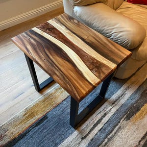 May include: A rectangular wooden side table with a natural wood grain top and black metal legs. The tabletop features a mix of light and dark brown wood tones. The table is approximately 30 inches long.