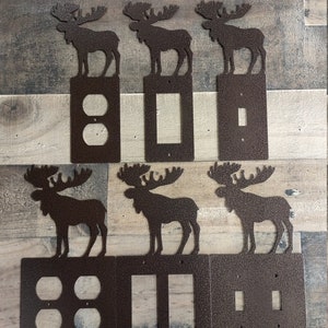 May include: Six metal light switch covers with moose silhouettes. The covers are in a rustic brown color and feature different switch configurations: a double outlet, a single switch, a double switch, a triple switch, and two single switches.