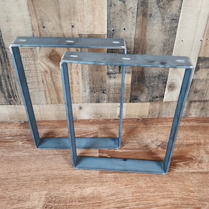 May include: Two metal table legs, one slightly larger than the other, are shown against a wood background. The legs are made of a dark gray metal and have a rectangular shape.
