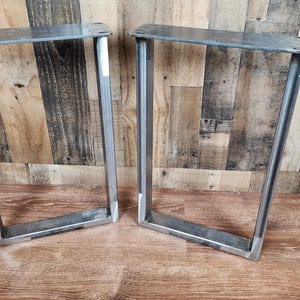 May include: Two silver metal table legs with a rectangular frame design. The legs are made of thick metal and have a welded construction. The legs are designed to be mounted to a tabletop. The legs are shown on a wooden surface.