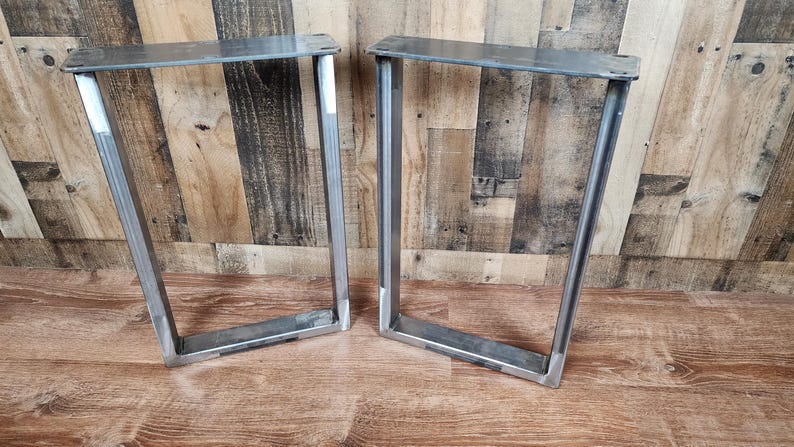May include: Two silver metal table legs with a rectangular frame design. The legs are made of thick metal and have a welded construction. The legs are designed to be mounted to a tabletop. The legs are shown on a wooden surface.