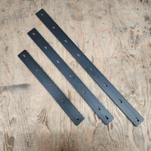 May include: Three black metal strips with multiple holes, arranged in a triangular pattern on a wood surface.