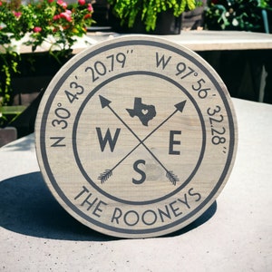 May include: A round wooden sign with a compass design featuring a black outline of the state of Texas with a heart in the center. The sign also includes the coordinates "N 30°43′ 20.79′ W 97°56′ 32.28" and the text "THE ROONEYS".