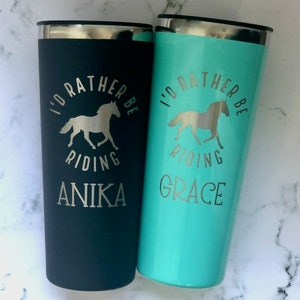 May include: Two stainless steel travel mugs with black and turquoise lids. The mugs are engraved with the text "I'd rather be riding" and a horse silhouette. The mugs are personalized with the names "Anika" and "Grace".