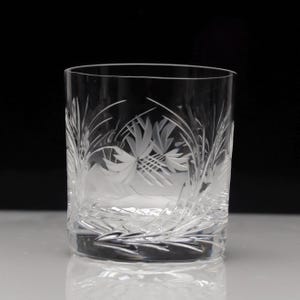 May include: A clear, etched glass tumbler with a botanical design. The glass features detailed carvings of leaves and floral patterns, creating a textured surface. The glass is cylindrical and sits on a reflective surface.