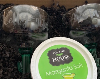 May include: Two clear glass margarita glasses with personalised names, "Kelly" and "Kristen", are nestled in black shredded paper. A white container with a green lid reads "On the House Margarita Salt" and features a margarita glass with a lime wedge.