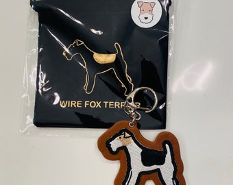 May include: A brown leather keychain featuring a white and black embroidered wire fox terrier design. The keychain is supplied with a black velvet pouch with a gold wire fox terrier silhouette and the text "WIRE FOX TERRIER".