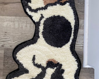 Puede incluir: Una alfombra con forma de perro con borde negro. La alfombra presenta un cuerpo blanco con una gran mancha negra y parches marrones en la cabeza. La alfombra está hecha de un material suave y texturizado.