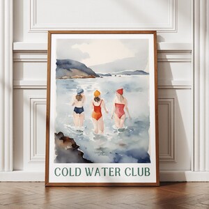 以下が含まれることがあります： 海に向かって歩く、水着と帽子をかぶった3人の女性を描いた水彩画。絵は茶色の木製の額縁に収められており、下部に「COLD WATER CLUB」の文字が書かれている。