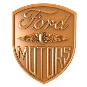 May include: A bronze-colored Ford Motors logo with a winged wheel in the center and the words "Ford Motors" in a stylized font.