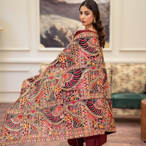 Peut inclure: Une femme portant une tenue marron avec un châle brodé coloré. Le châle présente un motif géométrique avec des miroirs et est drapé sur ses épaules.