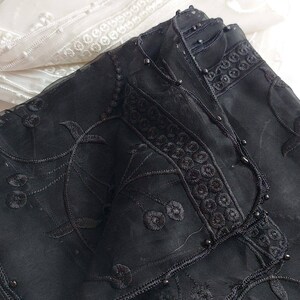 May include: Black sheer fabric with black embroidery and black beads. The fabric is folded and has a white sheer fabric underneath.