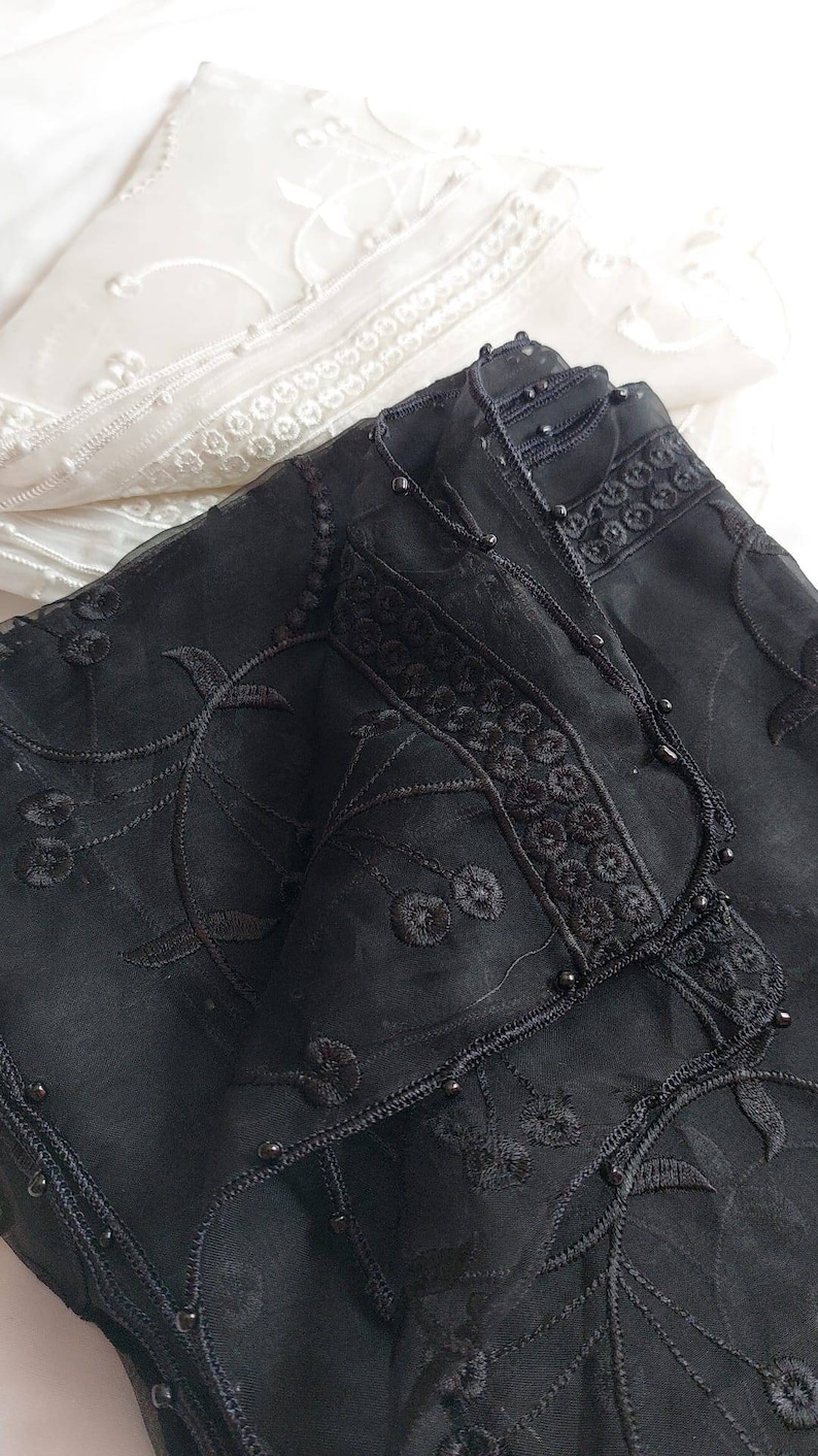 May include: Black sheer fabric with black embroidery and black beads. The fabric is folded and has a white sheer fabric underneath.