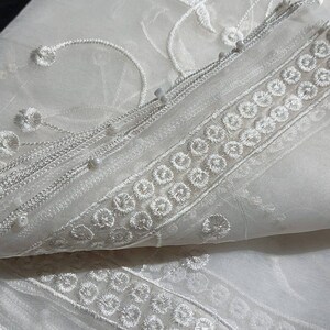 May include: White sheer fabric with delicate floral embroidery and a beaded trim. The fabric is folded in half, revealing the intricate details of the design.