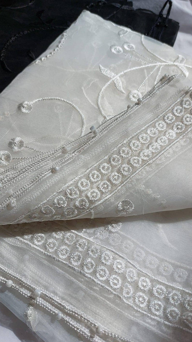 May include: White sheer fabric with delicate floral embroidery and a beaded trim. The fabric is folded in half, revealing the intricate details of the design.