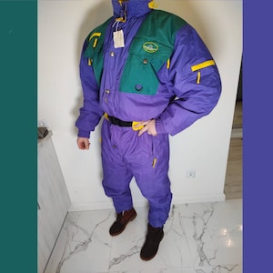 Może przedstawiać: Fioletowy, zielony i żółty kombinezon narciarski w stylu vintage. Jednoczęściowy kombinezon narciarski ma wysoki kołnierz, wiele kieszeni i czarny pasek. Kombinezon ma wzór w blokach kolorów z logo na piersi i żółtymi akcentami zamków błyskawicznych.