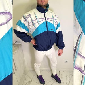 Puede incluir: Chaqueta deportiva vintage de estilo retro. La chaqueta presenta un diseño de bloques de color con paneles blancos, azul marino y turquesa. Tiene un cuello alto y una cremallera completa. La chaqueta tiene un patrón geométrico en morado y turquesa.