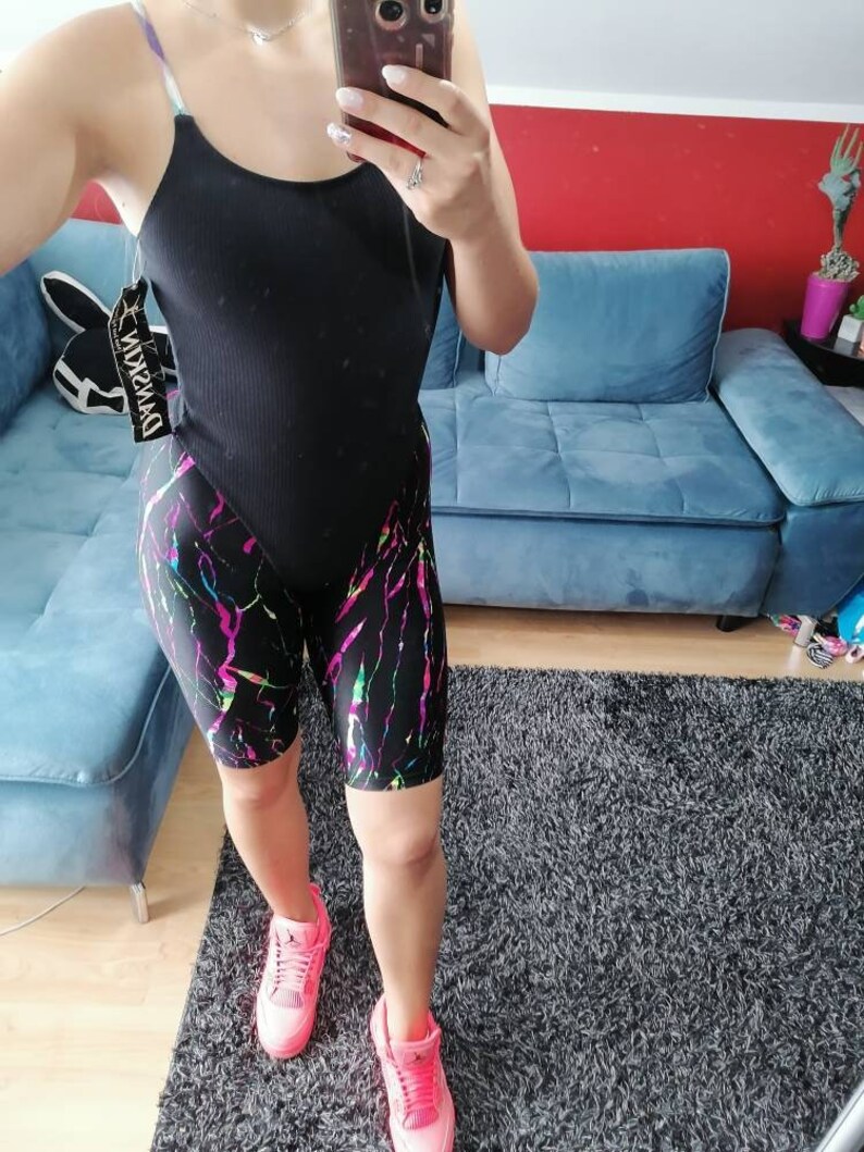 May include: A black bodysuit with a racerback style and a black and white patterned biker short with pink, green, and blue accents. The model is wearing pink sneakers.