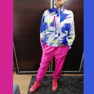 May include: A white, blue and pink tracksuit with a geometric print. The jacket has a zip closure and a high collar. The trousers have a relaxed fit. The model is wearing pink trainers.