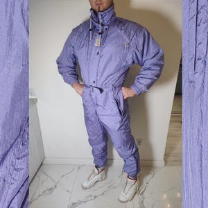 Może przedstawiać: Fioletowy kombinezon narciarski z zamkiem błyskawicznym z przodu i paskiem. Kombinezon ma kaptur i długie rękawy. Kombinezon jest wykonany z błyszczącego, nylonowego materiału.