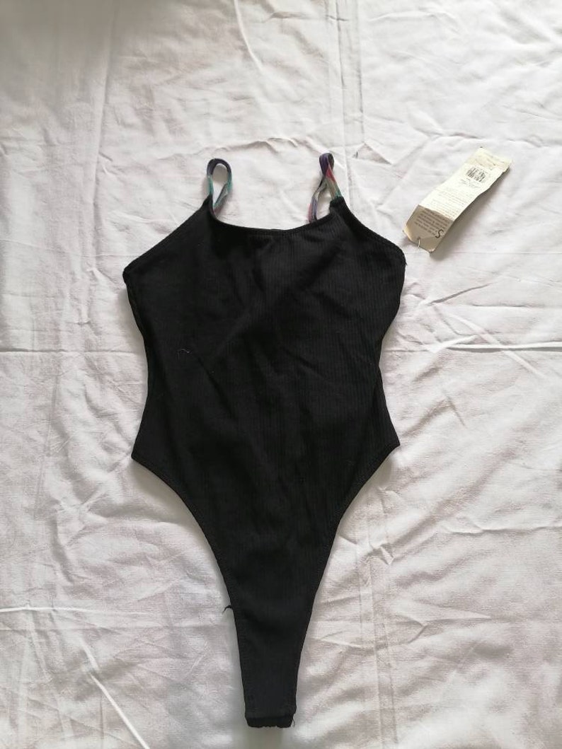 May include: Black bodysuit with thin rainbow straps. The straps are made of a woven fabric.