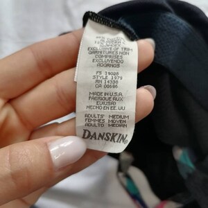 May include: A white tag with black text that reads "EXCLUSIVE OF TRIM GARNITURES NON COMPRISES EXCLUYENDO ADORNOS" and "MADE IN U.S.A. FABRIQUE AUX E.U.I.S.A. HECHO EN EE. UU" and "ADULTS MEDIUM FEMMES MOYEN ADULTO MEDIAN" with the Danskin logo at the bottom.