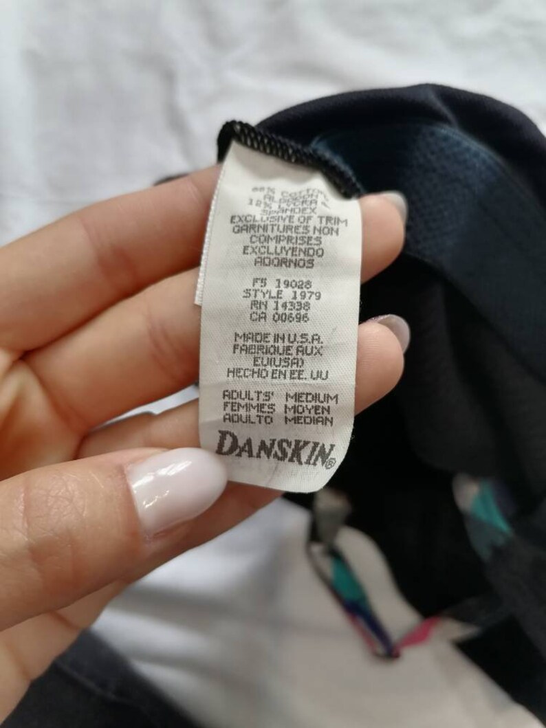 May include: A white tag with black text that reads "EXCLUSIVE OF TRIM GARNITURES NON COMPRISES EXCLUYENDO ADORNOS" and "MADE IN U.S.A. FABRIQUE AUX E.U.I.S.A. HECHO EN EE. UU" and "ADULTS MEDIUM FEMMES MOYEN ADULTO MEDIAN" with the Danskin logo at the bottom.