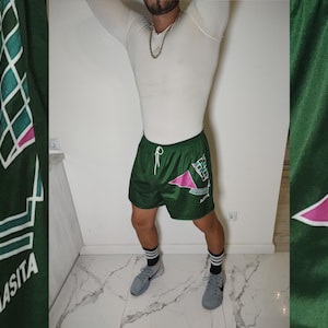 Peut inclure: Short de sport vert avec un motif géométrique rose et blanc. Le short a un cordon de serrage blanc et le mot "MASITA" est imprimé sur la jambe gauche.