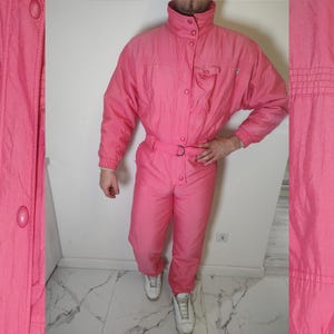 Może przedstawiać: Jasnoróżowy, vintage kombinezon narciarski. Jednoczęściowy kombinezon posiada wysoki kołnierz, zapinany na guziki przód i talię z paskiem. Kombinezon ma kieszeń na piersi i jest wykonany z błyszczącego, pikowanego materiału. Kombinezon narciarski jest zestawiony z białymi trampkami.