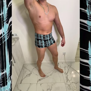 Puede incluir: Bañador a cuadros negros, turquesa y blancos. El bañador es corto y ajustado. El modelo está de pie frente a una pared blanca y un suelo de baldosas de mármol.