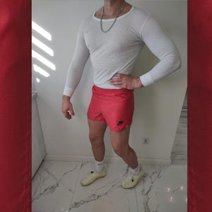 May include: A person wearing a white long-sleeve shirt and red athletic shorts with a black Nike logo. The person is also wearing white socks and light yellow shoes. A silver chain necklace is visible.