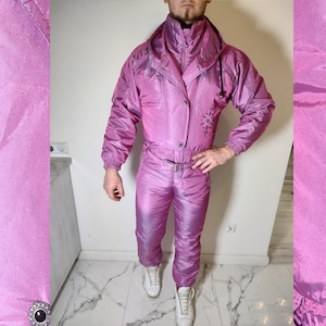 May include: A vintage, iridescent pink ski suit. The one-piece suit features a high collar, a zip-up front, and a belt at the waist. The suit has a starburst design on the chest and is paired with white sneakers. The fabric has a shiny, reflective quality.